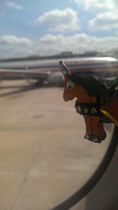A sparkle pony pin is held up to the window of a plane and is looking out at the tarmac. Another plane on the tarmac is in the background of the picture. The picture makes it seem as if sparkle pony is a bit sad to be flying home.