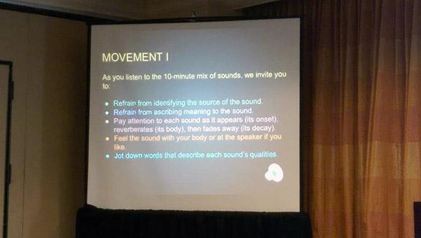 Image of a projected slide that reads: MOVEMENT 1: As you listen to the 10-minute mix of sounds, we invite you to: •Refrain from identifying the source of the sound. •Refrain from ascribing meaning to the sound. •Pay attention to each sound as it appears (its onset), reverberates (its body), then fades away (its decay). •Feel the sound with your body or at the speaker if you like. •Jot down words that describe each sound’s qualities.