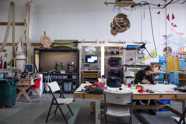 A man with fair skin and shoulder-length dark hair sits at a large table. He is hunched over small pieces of plastic, and the table is covered in plastic objects in a variety of shapes and colors. Along the wall behind him are large metal racks that hold 3D printers and computers.